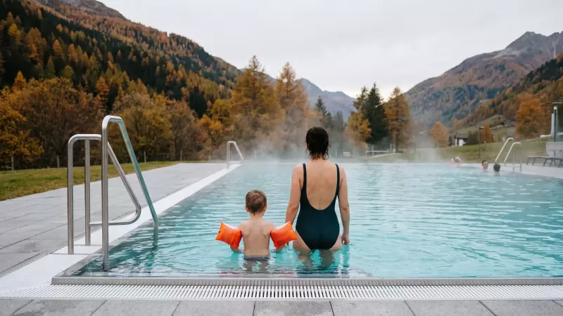 Parent et jeune enfant avec brassards, vus de dos dans un bassin thermal extérieur fumant au pied de montagnes d’automne, illustration IA