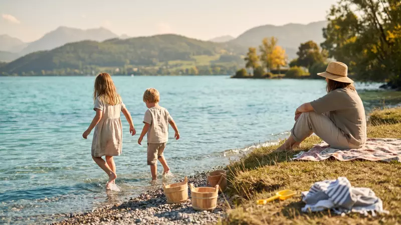 Deux jeunes enfants jouent pieds nus dans l’eau peu profonde au bord d’un lac suisse par un après-midi chaud de début septembre, un parent assis sur une serviette dans l’herbe derrière eux, montagnes au loin