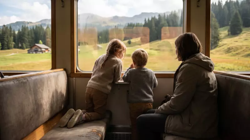 Deux enfants, vus de dos, observent un paysage de montagne de fin d’été depuis la fenêtre d’un train régional, un parent assis à côté d’eux