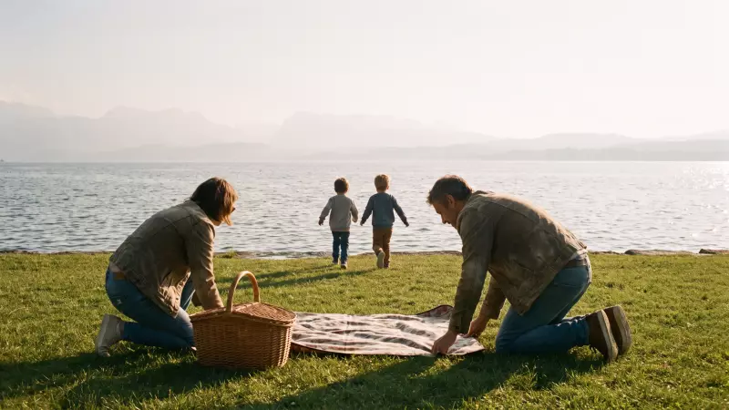 Une famille installe un pique-nique sur l’herbe au bord d’un lac suisse en fin d’après-midi, deux enfants courent vers l’eau